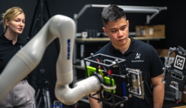 A man and a woman stand in a dark laboratory observing equipment. In front of them is a white robotic arm. The man's t-shirt has a logo for SpaceWERX.